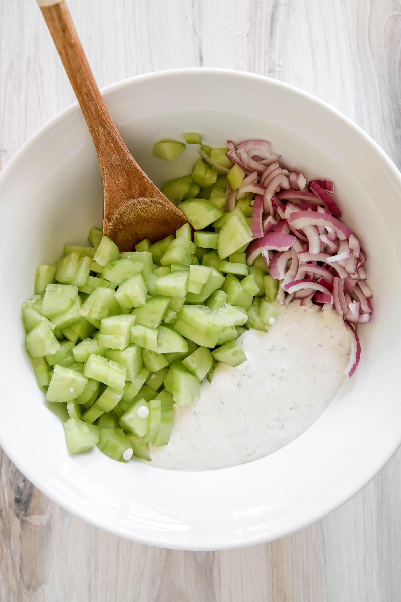 large white bowl with sliced cucumbers, diced red onions and creamy dressing.