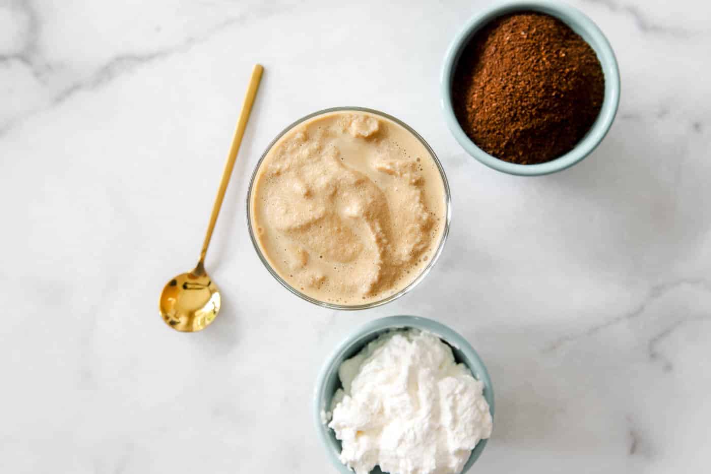 frosted coffee in a glass cup with whipped cream and espresso grounds nearby.